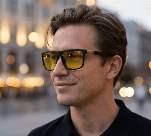 Mężczyzna w stylowych żółtych okularach do jazdy nocą, na tle rozmytych miejskich świateł, testuje lepszą nocną widoczność.