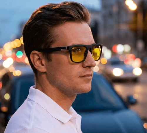 Młody mężczyzna w czarnych okularach z żółtymi soczewkami na tle rozmytych świateł wieczornego miasta.